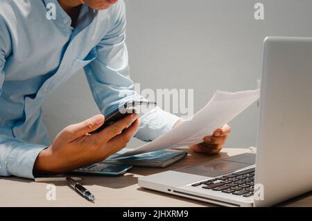 Uomo d'affari che tiene smartphone mobile e documenti cartacei finanza investimento e marketing concetto con computer portatile sulla scrivania. Foto Stock