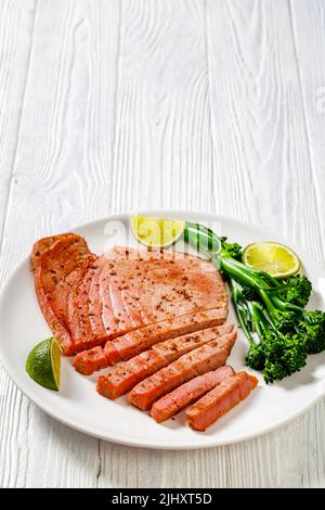 bistecca di tonno fritta con broccolini parboiled e lime su piatto bianco su tavola bianca in legno testurizzato, vista verticale dall'alto Foto Stock