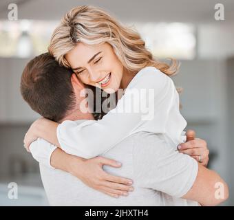 Bella donna che abbraccia il marito mentre sorride nella loro casa. Giovane moglie abbraccia il suo uomo mentre si trova in soggiorno. Sensazione di felicità e amore Foto Stock