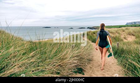 Donna surfista con muta e tavola da surf camminando lungo il percorso tra le dune verso la spiaggia Foto Stock