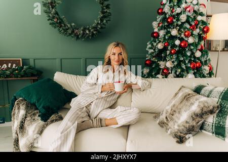 Una giovane donna europea in pigiama si siede su un divano la vigilia di Natale e beve caffè. Foto Stock