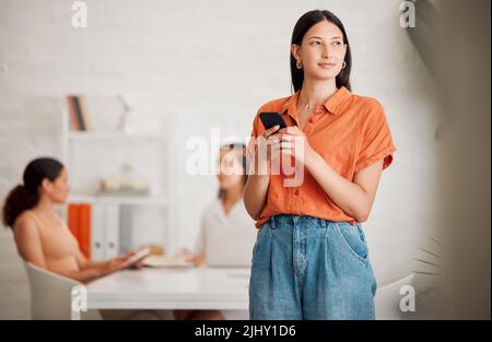 Una giovane donna ispanica che pensa mentre texting su un cellulare in un ufficio con i suoi colleghi in background. Imprenditore che fa un Foto Stock
