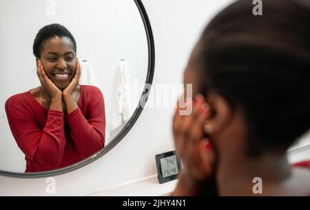 Ritratto di bella donna africana sorridente, guardando il suo volto in bagno specchio mani su guance. Parte della sua routine di cura di sé del mattino Foto Stock