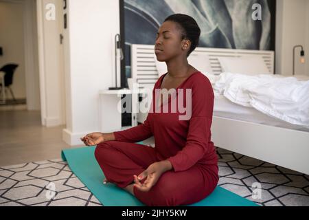 Bella donna africana nera seduta sul tappeto yoga al piano della camera da letto. Meditando nella posa del loto e gli occhi chiusi Foto Stock