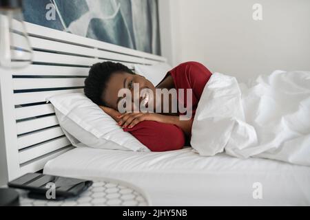 Bella donna africana che si stende a letto, guardando la macchina fotografica e sorridendo, adagiato comodamente sotto il piumone e sui cuscini al mattino, appena svegliato Foto Stock