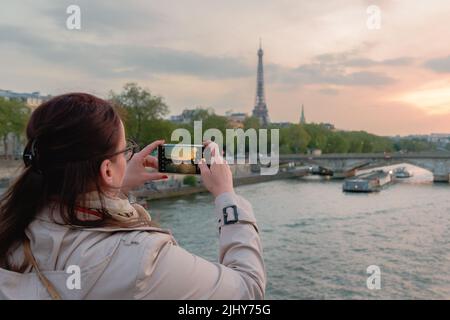 Giovane donna che fotografa con il cellulare dalla torre Eifel al tramonto. Con ponte e la Senna a Parigi. Foto Stock