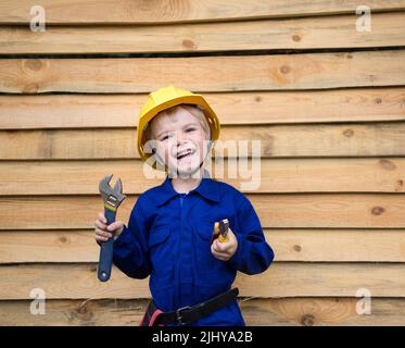 ritratto di un ragazzo grazioso inquisitivo di 6 anni in una uniforme blu tuta e in un casco da costruzione con attrezzi nelle sue mani su uno sfondo di legno. Foto Stock