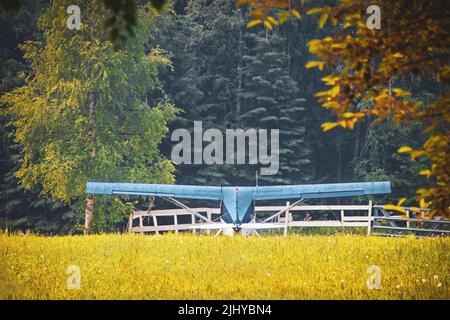 Piccolo aeroplano blu parcheggiato in campo vicino a recinto di legno bianco con sfondo sfocato della foresta sempreverde Foto Stock