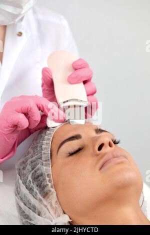 Cosmetologist, estetista in guanti rosa che fanno trattamento facciale con spatola ultrasonica a donna, trattamento di scrubber della pelle del viso con ultrasuoni Foto Stock