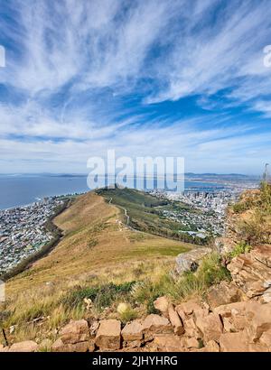 Montagna rocciosa con vista di una città costale sul mare a Città del Capo. Paesaggio di collina verde con pietre e piante circondate da una città urbana e. Foto Stock