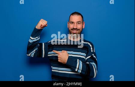 L'uomo barbuto spagnolo in pullover a strisce puntando il dito ai muscoli bicipiti dimostrando potenza di forza isolata su sfondo blu studio Foto Stock