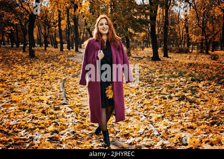 Ritratto autunnale all'aperto di sorridente felice più dimensione capelli rossi donna in cappotto a piedi nel parco autunnale. Foto Stock