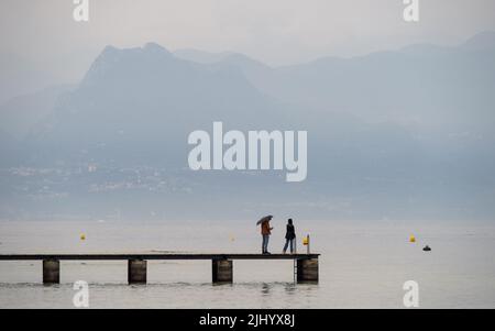 Un bel colpo di coppia in piedi su un molo in una giornata piovosa sul Lago di Garda, Italia Foto Stock