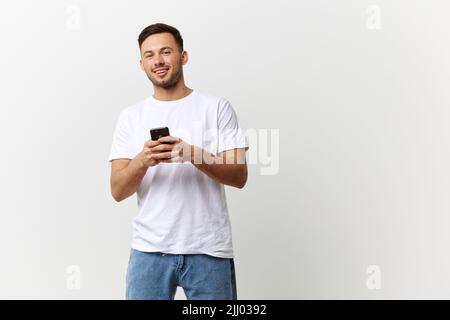 Allegro abbronzato bell'uomo in T-shirt di base digitando messaggio con il sorriso del telefono alla macchina fotografica che posa isolato su sfondo bianco studio. Spazio di copia Foto Stock