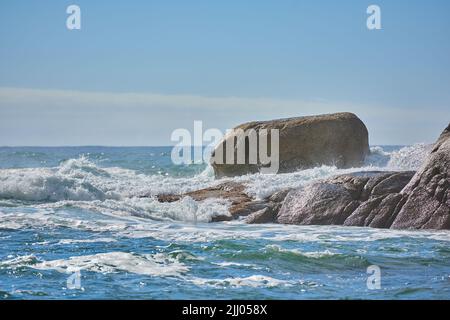 Copia lo spazio di un mare turbolento con maree ruvide e onde ondate da forti venti che si schiantano su grandi massi in spiaggia con un cielo blu chiaro Foto Stock