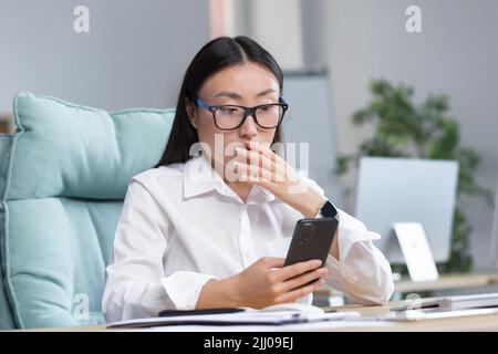 Preoccupata giovane bella donna asiatica d'affari in occhiali tiene il telefono nelle sue mani, guarda il telefono cellulare, ha ricevuto cattive notizie, sorpreso, scioccato. Seduta su una scrivania in un ufficio moderno. Foto Stock