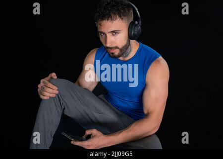 Bel maschio con tatuaggi nel suo 20s in palestra con telefono e cuffie, isolato su sfondo nero Foto Stock