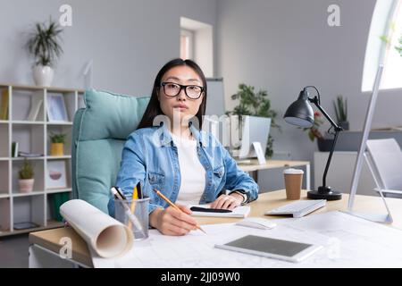 Ritratto di una giovane e bella donna asiatica architetto in occhiali che lavora a una scrivania a un computer e che si disegna di un progetto. Guarda nella fotocamera Foto Stock