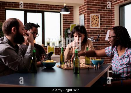 Gruppo multietnico intelligente di persone sedute a tavola mentre giocano a scacchi. Felici amici diversi seduti insieme a casa in soggiorno, godendo di giochi da tavolo strategia mentre si beve bevande e snack. Foto Stock