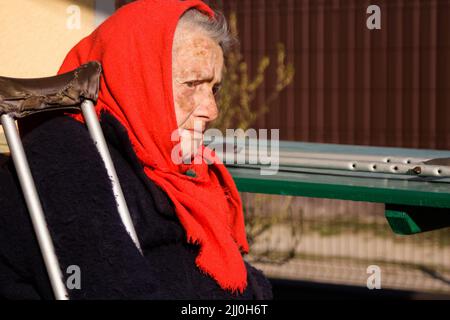 Defocus donna Ucraina anziana in scialle rosse con stampelle seduti all'esterno. Donna di 85 anni. Persona sola. Primo piano verticale. Malati e casalinga. Triste Foto Stock