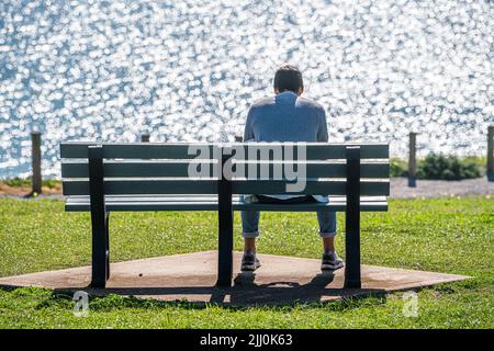 21 luglio 2022: Vista posteriore dell'uomo seduto su una panchina al sole di fronte all'oceano Foto Stock