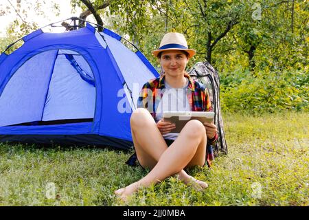 Defocus giovane donna che lavora sul tablet vicino campeggio tenda all'aperto circondato da bella natura. Freelance, sabbatico. Fuori fuoco. Foto Stock