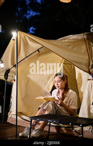 Tranquilla e rilassata giovane donna asiatica si siede di fronte alla sua tenda da campeggio, leggendo un libro di notte. Concetto di campeggio Foto Stock