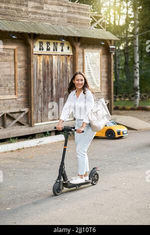 Giovane donna in elegante vestito ama guidare scooter elettrico sulla strada della città turistica. La signora bruna prova le nuove attività sulle vacanze estive Foto Stock