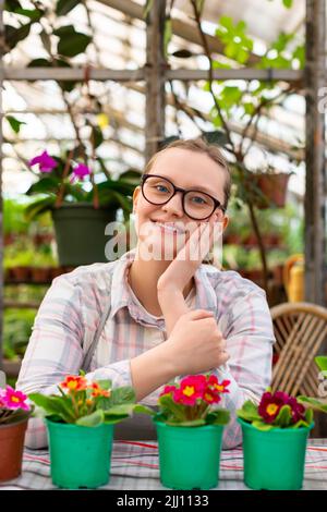 Una giovane donna con diversi fiori in pentole verdi in una serra che guarda la macchina fotografica e sorridente. Foto verticale. Foto Stock