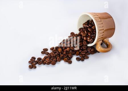 Caffè in grani in una tazza di caffè marrone isolato su bianco. I chicchi di caffè torrefatto fuoriescono dalla tazza di caffè capovolta con lo spazio di copia. Fuoco selettivo, vista laterale Foto Stock