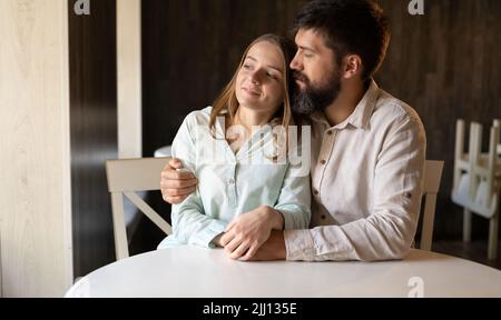 Giovane coppia seduta al tavolo da caffe' vicino alla finestra, uomo bearded che abbraccia moglie donna nel ristorante, datario e concetto romanticismo Foto Stock