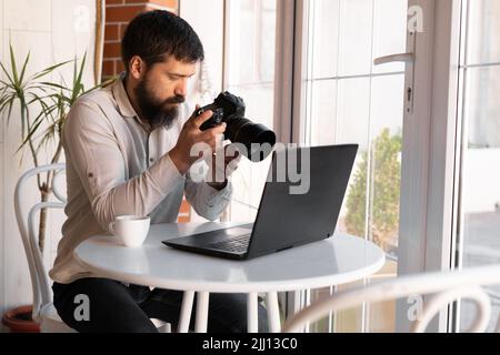 Fotografo creativo che lavora con laptop e fotocamera digitale al bar. Giornalista professionista o freelance che guarda le fotografie con fotocamera DSLR, c Foto Stock