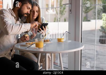 Giovane coppia al caffè, lei sta bevendo succo, sta mostrando qualcosa nel suo telefono cellulare. Sono felici e si sentono a proprio agio. Foto Stock