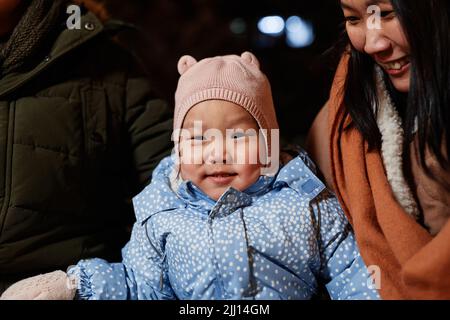 Ritratto medio di primo piano di bella ragazza asiatica toddler seduta con i suoi genitori all'aperto la sera d'inverno sorridendo alla macchina fotografica Foto Stock