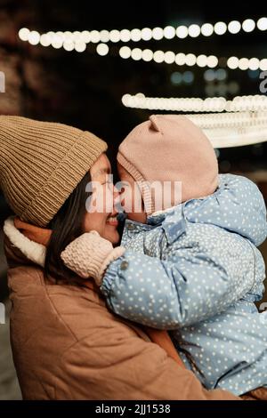 Colpo medio verticale della donna asiatica felice che tiene la sua figlia del toddler che passa il tempo grande all'aperto sulla sera di inverno Foto Stock