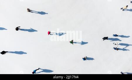 Molte persone stanno pattinando su una pista di ghiaccio bianca all'aperto in città in una soleggiata giornata invernale. Ombre di persone che pattinano sulla superficie di una pista di ghiaccio bianca. Vista aerea del drone. Vista dall'alto. Stile di vita, sport, riposo Foto Stock