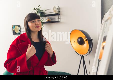 Donna caucasica dai capelli scuri con bangs e trucco morbido provando su un cappotto rosso in salotto guardando lo specchio. Scatto al coperto. Foto di alta qualità Foto Stock