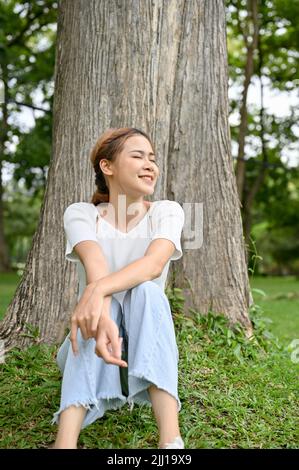 Bella e felice giovane donna asiatica si rilassa seduto sotto un albero nel bellissimo parco della città nel suo fine settimana. Foto Stock