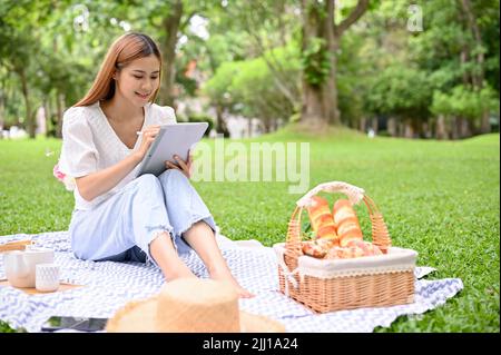 Attraente giovane donna asiatica usa un tablet portatile touchpad e si rilassa mentre si fa un picnic nel bellissimo parco verde. Foto Stock