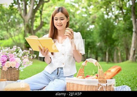 Bella giovane donna asiatica picking nel bellissimo parco della città, si rilassa leggendo un libro e mangiando muffin. Concetto di picnic Foto Stock