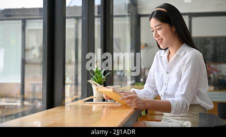 Attraente giovane donna asiatica si rilassa seduto nel caffè e godersi la lettura del suo libro preferito romanzo. Foto Stock