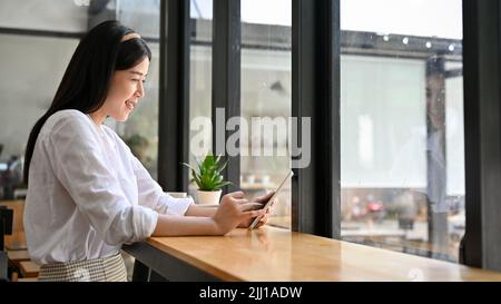 Bella giovane donna asiatica o studentessa college si rilassa seduto nel bar e utilizzando il touchpad portatile tablet. Foto Stock