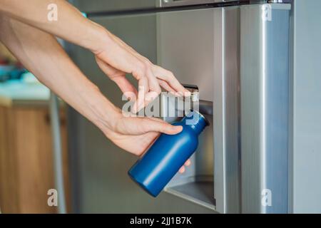 La mano maschio versa acqua fredda e cubetti di ghiaccio in una bottiglia di metallo dal distributore del frigorifero domestico Foto Stock