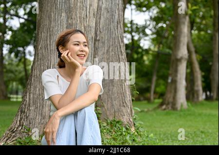 Attraente e felice giovane donna asiatica si rilassa seduto sotto un albero nella bella vegetazione di un parco. Foto Stock