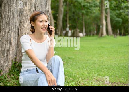 Bella giovane donna asiatica si rilassa mentre si siede sotto l'albero nel parco e al telefono chiamando qualcuno. Foto Stock