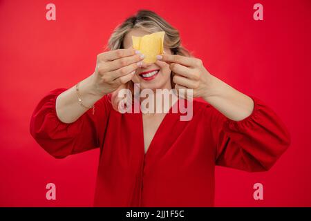 Donna bionda sorridente e gioiosa in abito rosso che tiene e copre gli occhi con patatine fritte, croccante nelle mani in studio rosso Foto Stock