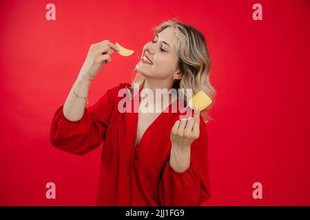 Sorridendo felice e gioiosa donna bionda in rosso vestito tenendo e mangiando patata chip, croccante nelle mani. Cibo spazzatura. Spazio di copia Foto Stock