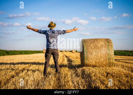 Coltivatore felice con le braccia allungate in piedi accanto alle balle di fieno dopo la raccolta riuscita. Foto Stock