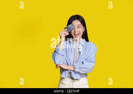 Ragazza asiatica eccitata sorridente coprila con l'occhio destro della scheda della banca pronto a spendere i soldi. Ragazza asiatica in camicia blu con carta mockup banca isolato su sfondo giallo. Concetto di shopping. Foto Stock