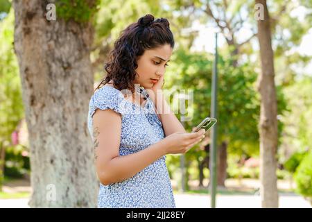 Vista laterale della giovane donna bruna che indossa un abito estivo sul parco cittadino, all'aperto tocca il mento e guarda lo schermo del telefono. Sembra infastidito e stressato Foto Stock